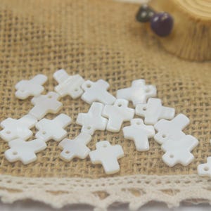 Peut inclure: Un ensemble de petits perles blanches en forme de croix, avec une finition brillante. Les perles sont dispersées sur une surface en toile de jute, avec une bordure en dentelle décorative. Les croix ont un petit trou pour l'enfilage.