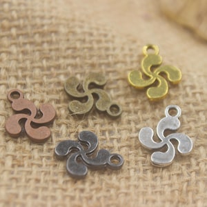May include: Five metal charms in various colors, including copper, bronze, gold, silver, and gunmetal. Each charm features a stylized, four-lobed design with a small loop for attaching to jewelry or other crafts. The charms are arranged on a textured, tan fabric.