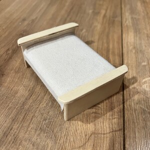 Könnte beinhalten: Ein Miniaturbett mit einem hellen Holzrahmen und einer hellgrauen Stoffmatratze. Das Bett hat ein schlichtes Design mit Kopf- und Fußteil. Das Bett steht auf einer Holzoberfläche.