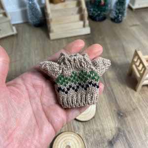 Könnte beinhalten: Ein Miniatur-Beige-Pullover mit grünen und schwarzen geometrischen Mustern. Das winzige Strickware hat lange Ärmel und wird in einer Hand gehalten. Der Hintergrund umfasst Miniatur-Weihnachtsbäume und eine kleine Holztür.