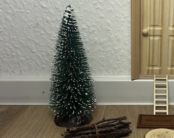 Könnte beinhalten: Eine kleine Holztür im Miniaturformat mit einer Willkommensmatte und einer kleinen Leiter, die zu ihr hinauf führt. Es gibt einen kleinen künstlichen Weihnachtsbaum mit weißem Schnee darauf, einen Stapel Stöcke und mehrere Holzscheiben auf dem Boden.