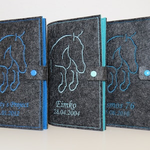 Könnte beinhalten: Drei dunkelgraue Filzhüllen für Notizbücher mit gestickten blauen Pferdesilhouetten und personalisiertem Text. Jede Hülle verfügt über einen passenden blauen Druckknopfverschluss und sichtbare Nähte an den Kanten sowie blaue Innenseiten.
