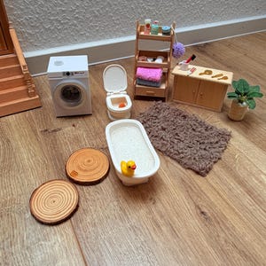 Könnte beinhalten: Ein Miniatur-Badezimmerset bestehend aus einer weißen Waschmaschine, einer Toilette, einer Badewanne mit gelber Quietscheente, einem Holzregal mit Handtüchern und Pflegeprodukten, einem Holzschrank, einem braunen Hochflorteppich und zwei Holzscheiben.