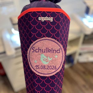 Könnte beinhalten: Eine violette, kegelförmige Schultüte mit rosa Fischschuppenmuster und leuchtend orangefarbenem Rand. Sie trägt den Schriftzug ergobag und einen runden Aufnäher mit dem Wort Schulkind, einem Meerjungfrauenmotiv, zwei Sternen und dem Datum 15.08.2026.