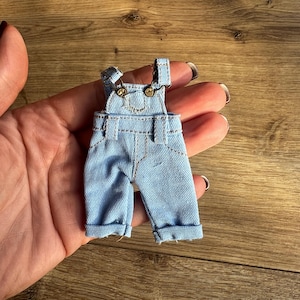 Könnte beinhalten: Hellblaue Jeans-Latzhose mit umgeschlagenen Säumen und messingfarbenen Beschlägen. Das Miniaturkleidungsstück hat verstellbare Träger und ein klassisches Latzhosendesign. Die Latzhose wird in einer Hand gehalten.