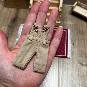 Könnte beinhalten: Miniatur-Latzhose in Beige mit Metallverschlüssen, einschließlich Hosenträgerclips und Knöpfen. Das kleine Kleidungsstück besteht aus einem strukturierten Stoff und hat umgeschlagene Manschetten. Die Latzhose ist für eine Puppe oder eine Miniaturfigur konzipiert.