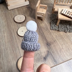 Könnte beinhalten: Eine graue Strickmütze mit einem weißen Bommel oben. Die Mütze wird auf einem Finger getragen.