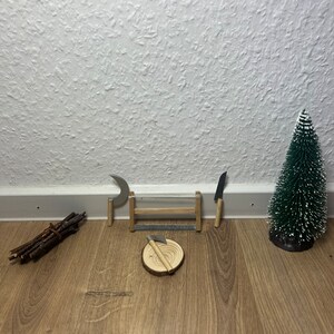 Może przedstawiać: Miniaturowa drewniana chatka z małym toporem, piłą, p&oacute;łksiężycem i stosami patyk&oacute;w na drewnianej podłodze. W tle znajduje się małe sztuczne drzewko sosnowe.