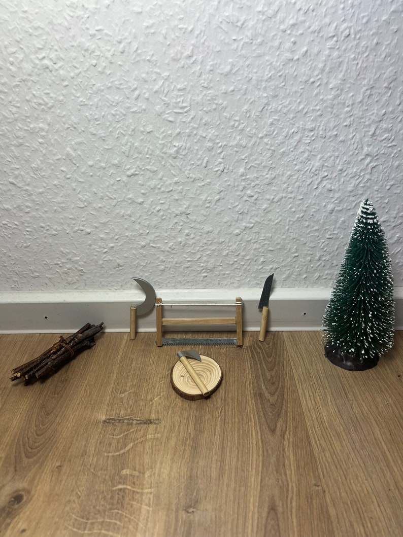 Może przedstawiać: Miniaturowa drewniana chatka z małym toporem, piłą, p&oacute;łksiężycem i stosami patyk&oacute;w na drewnianej podłodze. W tle znajduje się małe sztuczne drzewko sosnowe.