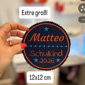 Könnte beinhalten: Ein kreisförmiger, dunkelblauer Stickaufnäher mit gezacktem Rand, der den orangefarbenen Text 'Matteo', 'Schulkind' und '2026' zeigt, akzentuiert durch hellblaue Sterne und eine gepunktete Linie. Der Aufnäher misst 12x12 cm und enthält 'Extra groß!'.