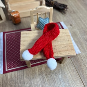 Könnte beinhalten: Ein miniaturer, rot gestrickter Schal mit weißen Bommeln an jedem Ende liegt auf einem kleinen Holztisch. Der Schal ist Teil einer Puppenhaus-Szene, mit einem Miniaturstuhl, einem Teppich und anderen Möbeln im Hintergrund.