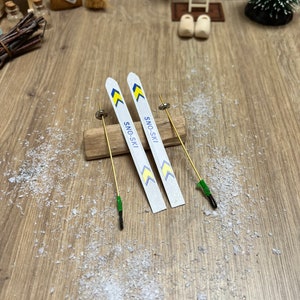 Könnte beinhalten: Ein Paar Miniatur-Skier in Weiß mit gelben und blauen Akzenten. Die Skier sind mit "SNO-SKI" beschriftet und liegen auf einer Holzoberfläche mit einer goldenen Metallstange.