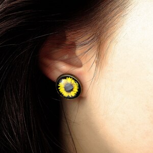 May include: A close-up of a person's ear with a sunflower earring. The earring is a round, gold-colored metal setting with a glass cabochon featuring a yellow sunflower with a black center.