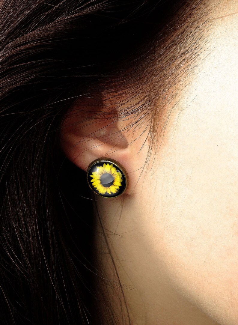 May include: A close-up of a person's ear with a sunflower earring. The earring is a round, gold-colored metal setting with a glass cabochon featuring a yellow sunflower with a black center.