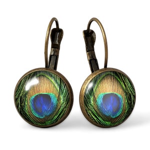 Puede incluir: Un par de pendientes de aro de bronce con un dije redondo de vidrio que presenta un diseño de pluma de pavo real. La pluma es verde, azul y dorada con un ojo azul en el centro.