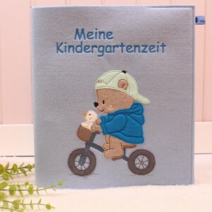Könnte beinhalten: Ein hellblauer Filz-Buchumschlag mit dem gestickten Text "Meine Kindergartenzeit". Der Umschlag zeigt einen gestickten Bären mit Mütze und blauer Jacke, der mit einer Ente im Korb Fahrrad fährt.