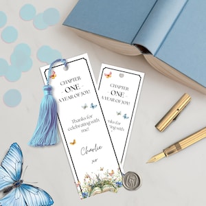 May include: Two white bookmarks with black borders, featuring text and floral designs. The text reads "CHAPTER ONE - A YEAR OF JOY!" and "Thanks for celebrating with me!" A blue tassel and a butterfly are attached to one bookmark. A gold pen and a blue book are also visible.