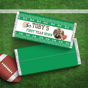 May include: Green and white candy bar wrapper with a football theme. It says "TOBY'S FIRST YEAR DOWN" and has a photo of a baby boy wearing a football helmet and holding a football. The wrapper has a ruler design with markings from 1 to 50.