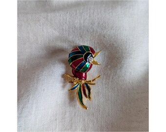 May include: A gold-tone brooch in the shape of a bird with red, green, and blue enamel. The bird has a black head with a single rhinestone eye and an open beak.