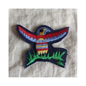 May include: A blue, red, yellow, and green embroidered patch of a stylized bird with outstretched wings, perched on a green leafy branch.