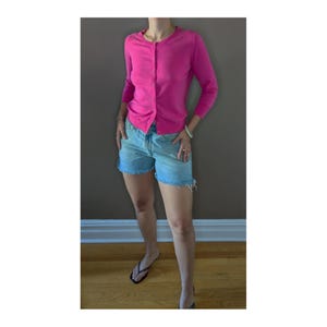 May include: A bright pink cardigan with three-quarter length sleeves and a round neckline, featuring a vertical row of small buttons down the front. The garment is paired with light blue denim shorts that have a frayed, raw-edge hemline.