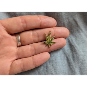 May include: A gold-coloured pin with a green marijuana leaf design.