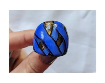 Peut inclure: Une bague bleue et dorée avec un motif géométrique. La bague est en verre et a une finition brillante.
