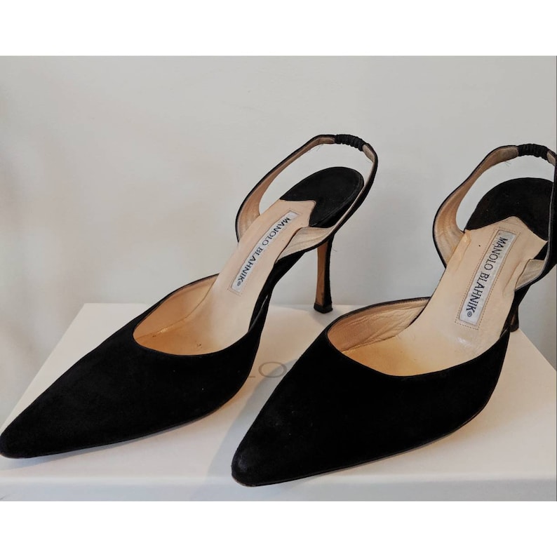May include: A pair of black suede heeled slingback shoes with a pointed toe. The shoes have a thin stiletto heel and a slingback strap that is secured with a small buckle. The shoes are labeled "Manolo Blahnik" on the insole.