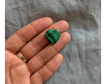 May include: A heart-shaped pendant made of green malachite with dark green veining. The pendant has a small gold-colored loop at the top for attaching to a chain or cord. The pendant is held in a hand against a gray background.