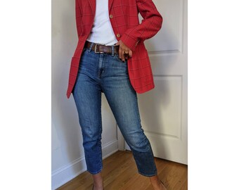 Op de afbeelding: Een rood geruit blazer met een bruine leren riem en een blauwe jeans. De blazer heeft een dubbele knoopsluiting en een revers.