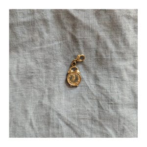 May include: A gold-colored charm in the shape of a small alarm clock. The clock face has black numbers and hands, and the charm has a clasp for attaching to a bracelet or necklace. The charm is on a light blue fabric.