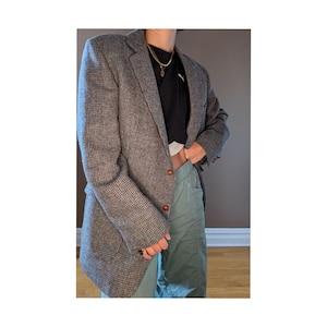 May include: A gray and black tweed blazer with a single button closure. The blazer has a brown button and a brown lining. The blazer is worn over a black shirt and green pants.