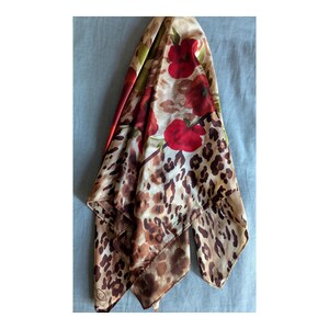 May include: A square scarf featuring a bold leopard print pattern in shades of brown, tan, and black, accented with large, vibrant red floral motifs and green leaves. The fabric has a smooth, satin-like finish with visible folds and a small logo tag.