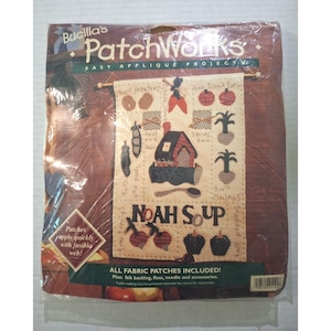 May include: A Bucilla PatchWorks easy applique project kit for a wall hanging titled Noah Soup. The package contains fabric patches, felt backing, floss, and a needle to create a cream-colored banner featuring a house, vegetables, and text.