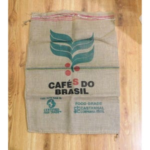 May include: A rectangular tan jute burlap sack featuring a green leaf graphic and red berries. The text reads CAFÉS DO BRASIL, THIS JUTE BAG IS FOOD GRADE, CERTIFIED FAIR TRADE, and CASTANHAL COMPANHIA TEXTIL. The top edge has a red and white stitched line.