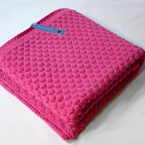 Op de afbeelding: Een roze gebreide babydeken met een textuurpatroon. De deken is gevouwen en heeft een label met de tekst "Handarbeit".