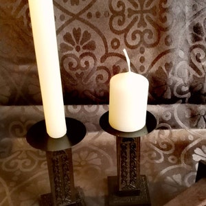 Könnte beinhalten: Zwei schwarze Kerzenhalter aus Metall mit verzierten Designs. Der größere Halter hat eine hohe weiße Kerze, während der kleinere Halter eine kürzere weiße Kerze hat.