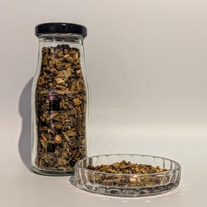 Könnte beinhalten: Ein Glasgefäß mit schwarzem Deckel, gefüllt mit getrockneten Kräutern, und eine kleine Glasschale mit getrockneten Kräutern.