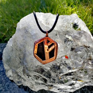 Op de afbeelding: Een koperen ketting met een zeshoekige hanger met een runesymbool erin. De hanger hangt aan een zwart koord en ligt op een helder kwartskristal.