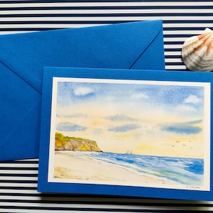 Könnte beinhalten: Eine blaue Grußkarte mit einem Aquarellgemälde einer Strandszene, einem passenden blauen Umschlag und einer Muschel auf gestreiftem Hintergrund. Das Gemälde zeigt einen Sandstrand, Meereswellen und einen bewölkten Himmel.