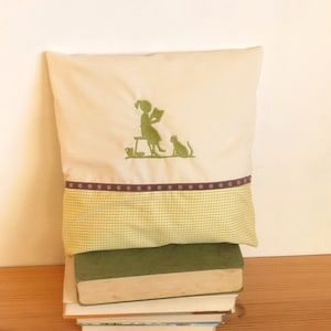 Könnte beinhalten: Ein weißes Kissen mit einem grünen gestickten Design einer Lesenden mit einer Katze, die neben ihr sitzt. Das Kissen hat ein grünes und weißes kariertes Muster auf der Unterseite und ein braunes Band mit weißen Punkten.