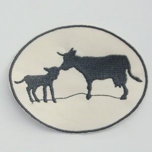 Könnte beinhalten: Ein weißer runder Patch mit einem gestickten Motiv einer schwarzen Kuh und eines Kalbes. Die Kuh leckt dem Kalb den Kopf.