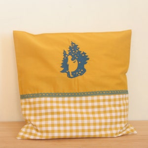 Könnte beinhalten: Senfgelbes Kissen mit einer blauen Silhouette einer Person und Bäumen. Das Kissen hat unten ein Vichy-Muster und eine türkisfarbene Spitzenborte. Das Kissen ist aus Stoff.