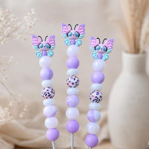Op de afbeelding: Drie decoratieve spiesen met vlinderopzetstukken. Elke spies heeft een vlinder met paarse, blauwe en witte accenten en een reeks paarse, witte en bedrukte kralen. De spiesen staan op een licht houten oppervlak.
