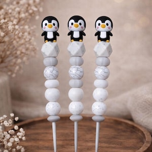 Könnte beinhalten: Drei Häkelnadeln mit einem dekorativen Aufsatz in Form eines schwarz-weißen Pinguins mit orangefarbenem Schnabel. Jeder Griff ist mit einer Reihe von Perlen in Weiß-, Grau- und Marmortönen verziert, die vertikal über der Nadel angeordnet sind.