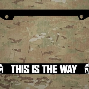 May include: Black license plate frame with white text that reads "THIS IS THE WAY" and two white Mandalorian helmet silhouettes on either side.
