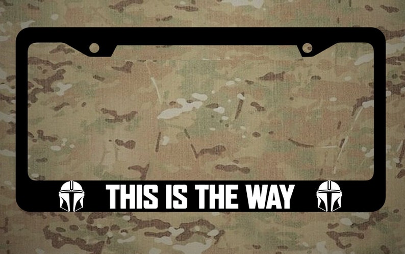 May include: Black license plate frame with white text that reads "THIS IS THE WAY" and two white Mandalorian helmet silhouettes on either side.