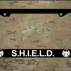 May include: Black license plate frame with white text that reads "S.H.I.E.L.D." and two white logos on either side of the text.