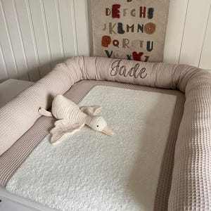 Può includere: Un tappetino per il cambio del bambino rosa chiaro e bianco con una superficie bianca e soffice e un bordo rosa chiaro con una trama a nido d'ape. Il tappetino ha un giocattolo di peluche a forma di oca bianca sopra e un'appendice da parete con l'alfabeto in lettere colorate e il nome "Jade" ricamato sotto.