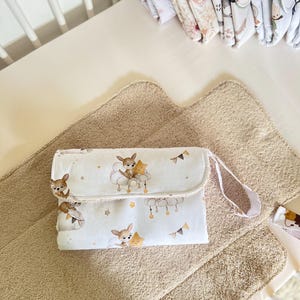 May include: A white changing pad with a cute deer and star print. The pad has a brown strap and is folded in half. It is sitting on a brown, fluffy changing mat.